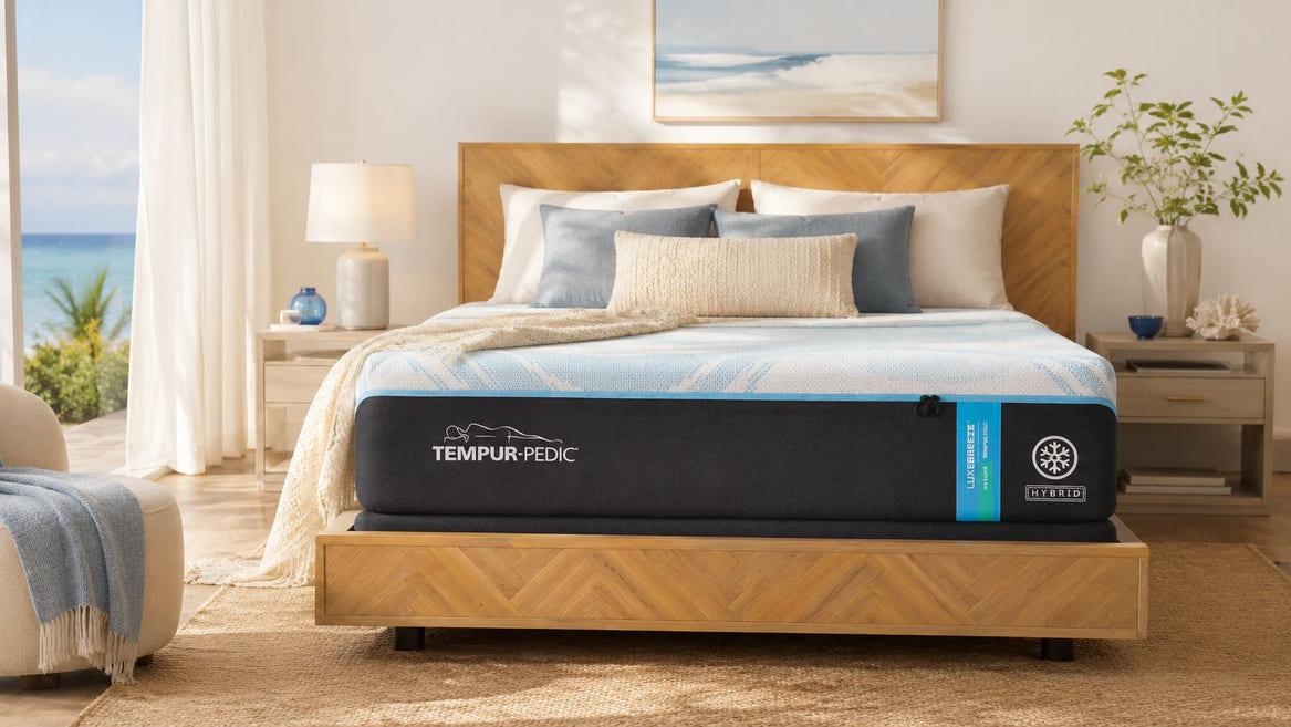 A TEMPUR-PEDIC Cloudbed mattress in black and blue is shown on a wooden bed frame in a beautifully styled bedroom with a view of the ocean through the window.